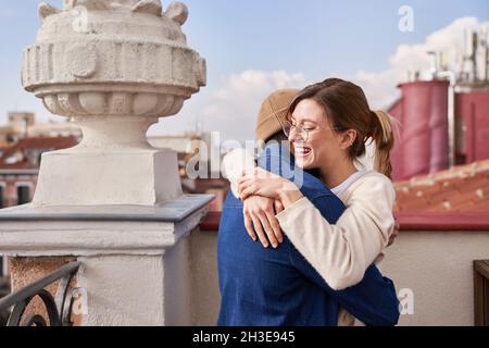 Side view of young girlfriend hugging boyfriend smiling brightly while standing with glass of juice on balcony Stock Photo