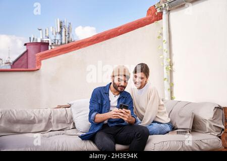 Smiling young Woman sitting close to bearded boyfriend and browsing smartphone on couch on terrace Stock Photo