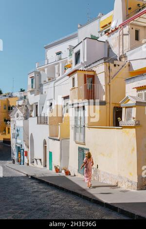 Back view of unrecognizable female in dress walking on asphalt walkway on street near aged buildings with colorful walls in town on Procida Island in Stock Photo