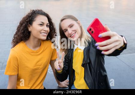From above of happy young multiethnic female friends smiling and showing two fingers gesture while taking selfie on street Stock Photo