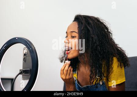 African American female applying lipstick while looking at camera on cellphone placed on ring light Stock Photo