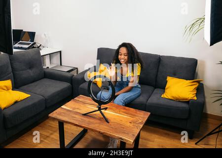 From above of ethnic Woman sitting on couch and greeting audience while recording vlog for smartphone placed on ring lamp Stock Photo