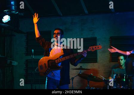 Young man playing bass guitar while performing in light club with neon illumination waving hand to public Stock Photo
