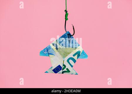Fish shaped banknote hanging on metal hook with green rope against pink background in light modern studio Stock Photo