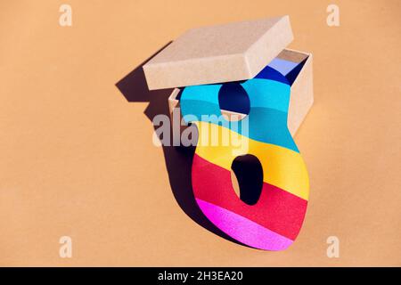 Multicolored masquerade mask for festive event placed in opened cardboard box with lid on orange background in light modern studio Stock Photo