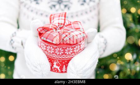Giving gifts for Christmas. The girl in knitted sweater holds out a gift box against the background of fir trees and golden bokeh. Red round gift box Stock Photo