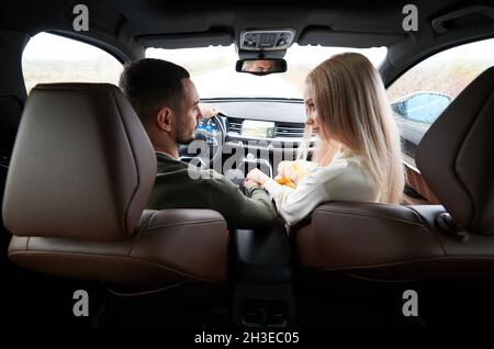 Happy man and woman traveling in car. Back view of beautiful young couple sitting on front passenger seats and smiling while handsome man driving comfortable automobile. Stock Photo