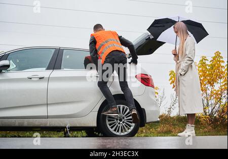 Elegant young woman standing by the car and holding umbrella while auto mechanic unscrewing lug nuts on wheel. Young man in vest repairing woman car on the road. Concept of emergency road service. Stock Photo