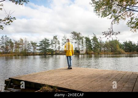 Woman wearing a yellow coat standing on a wooden pier by a lake looking at the view. Stock Photo