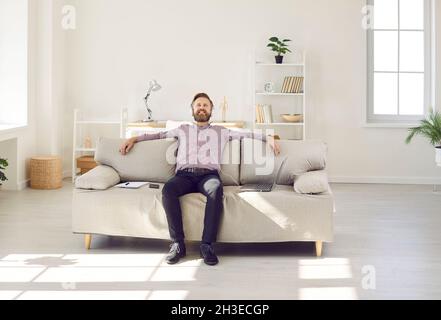 Happy man sitting on couch at home, listening to music through headphones and relaxing Stock Photo