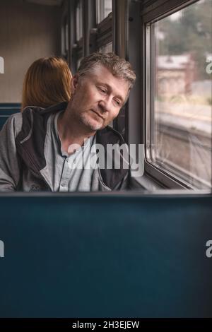 A serious handsome middle-aged man is sitting in the carriage of an old electric train and thoughtfully looks out the window. Work in another city. Stock Photo
