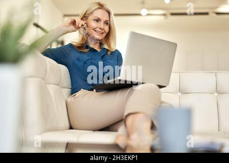 Cheerful mature businesswoman working from home, having online video call using laptop while sitting on a couch in modern apartment Stock Photo