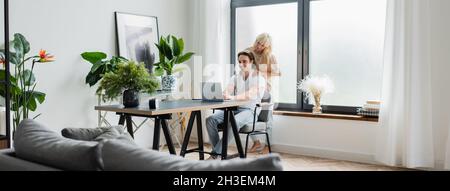 blonde woman massaging shoulders of boyfriend working from home with laptop, banner Stock Photo