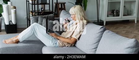 blonde woman drinking red wine while using laptop in living room, banner Stock Photo