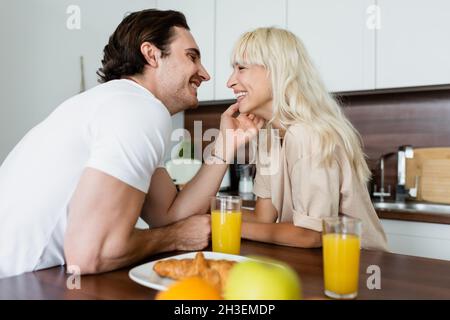 tattooed man touching face of happy girlfriend near glasses with orange juice and croissant Stock Photo