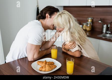 tattooed man touching face of happy girlfriend near glasses with orange juice and croissants on plate Stock Photo