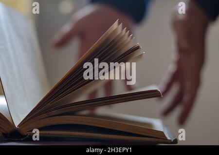 A defocused shot of an old open book and two open male hands. Palms in the background. Pages of hardcover book. Side view. Inside the room. Selective Stock Photo