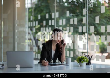 Smiling young asian businesswoman sit at desk smiles talking on smartphone looking out the window enjoy pleasant conversation with client, female Stock Photo