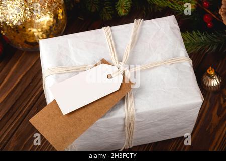 Christmas Gift Tag mockup. White minimal gift box on wooden table with festive decorations. Stock Photo