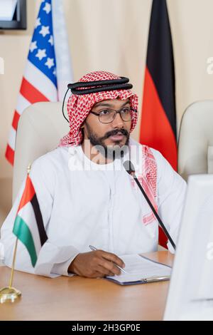 Serious Islamic politician in national dress sitting at table with papers and speaking into microphone at conference Stock Photo