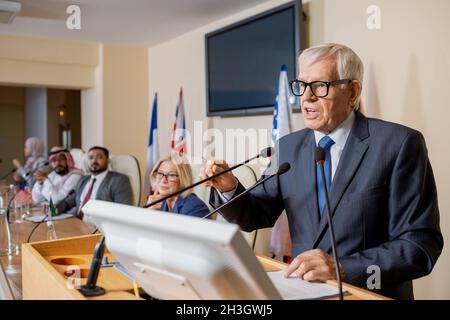 Emotional senior white-haired politician in glasses and suit gesturing hand while giving inspirational speech at press conference
