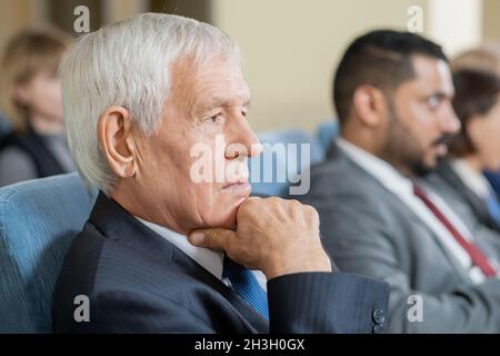 Pensive senior man sitting in armchair and touching chin while listening to speaker at business seminar Stock Photo