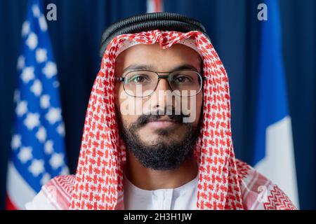 Portrait of serious confident Arabian politician in red and white ghutrah wearing agal and eyeglasses against national flags Stock Photo