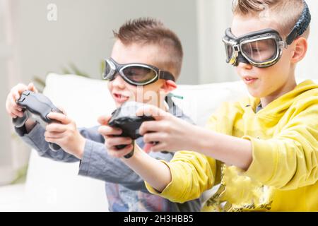 Two competitive young boys playing computer games seated side by side on a sofa with raised games controllers wearing goggles Stock Photo
