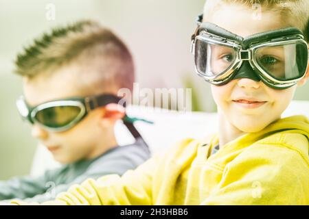 Cute high key portrait of a young boy wearing goggles turning to grin at the camera as he relaxes with his brother or friend on a sofa Stock Photo