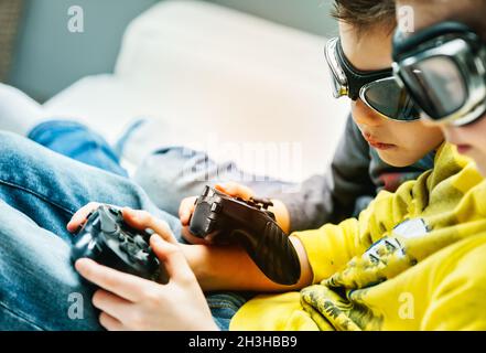 Young brothers wearing goggles huddling together playing video games on a sofa in a close up cropped view with focus to their hands and the controller Stock Photo