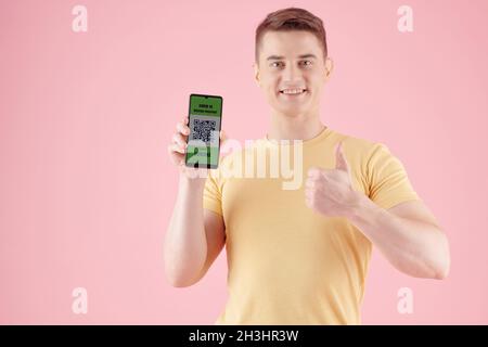 Portrait of excited smiling young man showing thumbs-up and smartphone with QR code after getting vaccinated Stock Photo