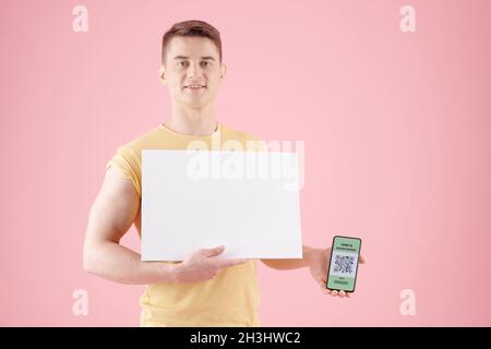 Positive handsome young man holding white sheet of paper and smartphone with vaccine passport Stock Photo
