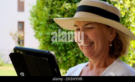 smiling Senior female blonde businesswoman in hat uses tablet pc, digital tablet for business work or study in her own green garden. woman aged 50-55 Stock Photo