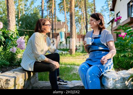 Two middle aged women sitting outdoors together in garden, backyard with mugs of tea Stock Photo