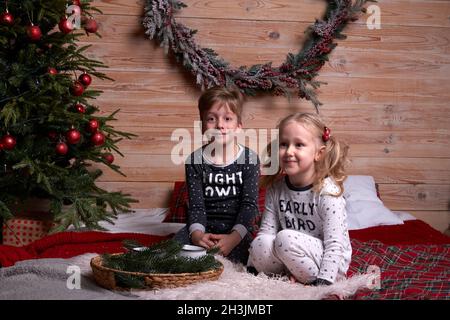Children in the same family look soft warm pajamas playing at home in christmas evening under decorated Christmas tree. Happy kid jumping on the bed. Stock Photo
