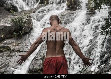 Back view of muscular man standing with raised arms near waterfall Stock Photo