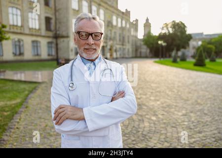 Portrait of confident senior male doctor in lab coat and glasses smiling at camera, standing with arms crossed outdoors near clinic Stock Photo