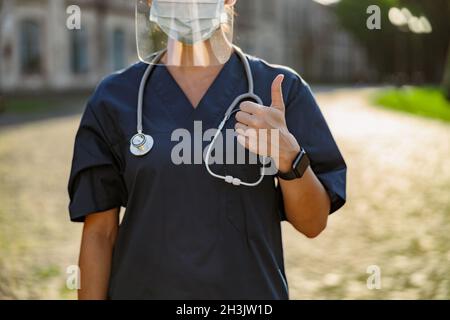 Cropped shot of young woman nurse in uniform wearing face shield and mask for safety to prevent Covid19, showing thumbs up, standing outdoors on a Stock Photo
