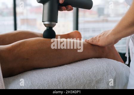 Close-up side view of unrecognizable professional male masseur massaging leg calf muscles using massage gun percussion tool of muscular athlete man. Stock Photo