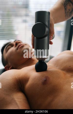 Medium shot of unrecognizable professional male masseur receiving chest muscles using massage gun percussion tool of muscular athlete man Stock Photo