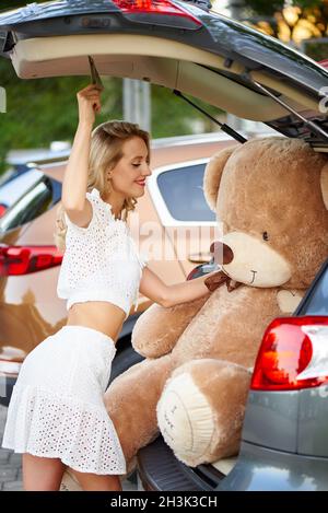 Happy young woman put a huge teddy bear in the car. Stock Photo