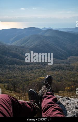 Human traveler sits on top of cliff and stretches feet in boots forward, enjoying views of autumn forest and mountain peaks. First person view. Travel Stock Photo