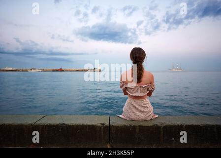 The back view of a lonely young woman sitting by the ocean at daylight. Stock Photo