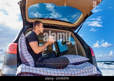 A young man with a cup of hot tea is resting in the car on the shore of the ocean. Stock Photo
