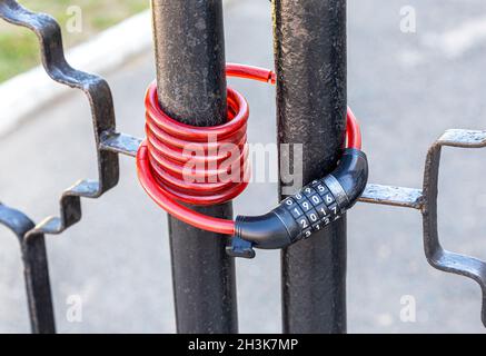Flexible red bicycle lock with the code set hangs on metal gates, close up. Security and safety concept. Stock Photo
