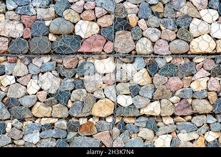 Stone wall with metal grid and rough stones as background. Rock texture in metal grid. Gabion net with stones Stock Photo