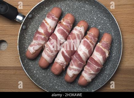 Preparation of raw sausages wrapped spirally in bacon on a frying pan. Top view. Stock Photo