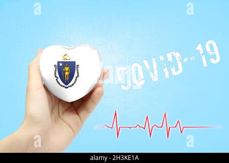 A girl is holding a toy in the shape of a heart with the flag of State of Massachusetts, a concept of health care during the covid-19 coronavirus pand Stock Photo