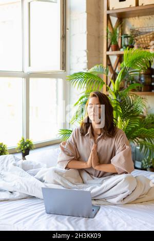 Young adult woman meditating in bed in front of laptop monitor. The concept of restoring strength and mental energy through yoga practice. Vertical ph Stock Photo