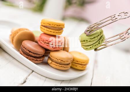 Picking a pistachio flavored macaron with pastry tongs from a selection of cookies on a plate, tilted view Stock Photo